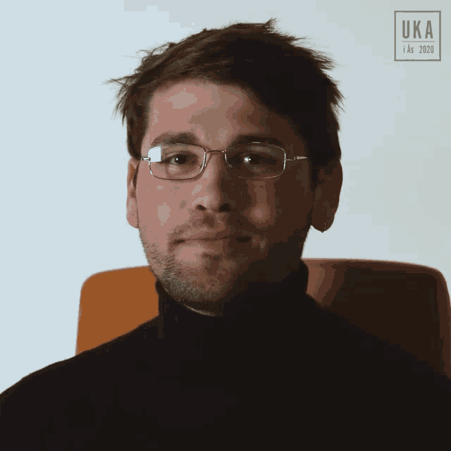 a man wearing glasses and a black turtleneck is sitting in a chair with a uk a logo behind him