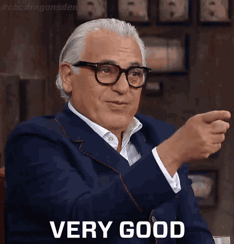 a man in a suit and glasses says very good while pointing at something