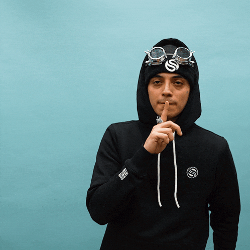 a man wearing a hoodie and goggles holds his finger to his mouth