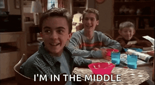 a boy is sitting at a table with two other boys and says `` i 'm in the middle ''