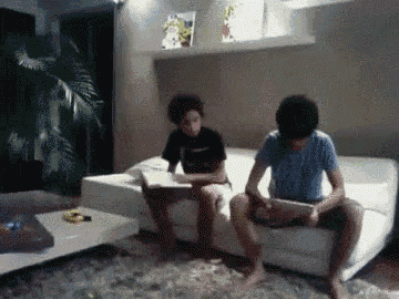 two boys are sitting on a couch in a living room reading books