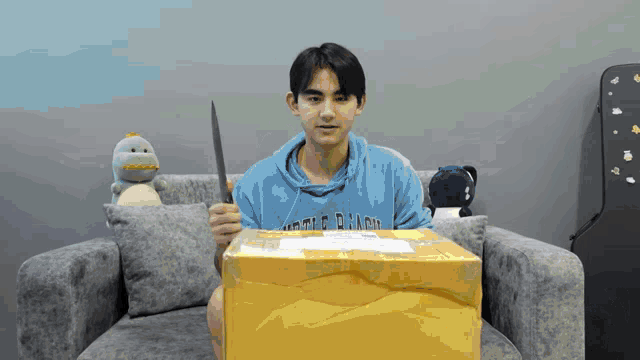a young man in a blue hoodie holds a knife in front of a cardboard box that says little dragon on it