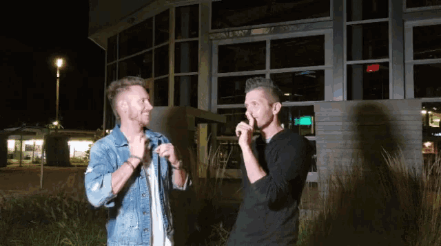 two men are standing in front of a building and one is covering his mouth with his hand