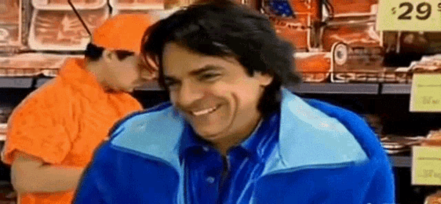 a man in a blue jacket is smiling in a grocery store while a man in an orange shirt is standing behind him .