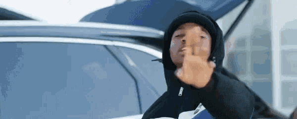 a man in a black hoodie is giving the middle finger while standing in front of a car .