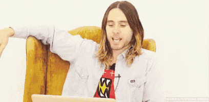 a man with long hair is sitting in a chair with his arms outstretched and a laptop in front of him .