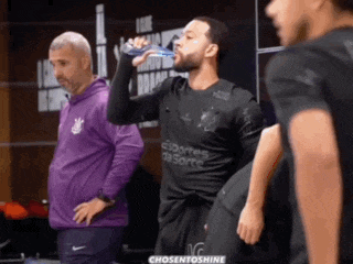 a man drinking water from a bottle while standing next to a man in a purple jacket