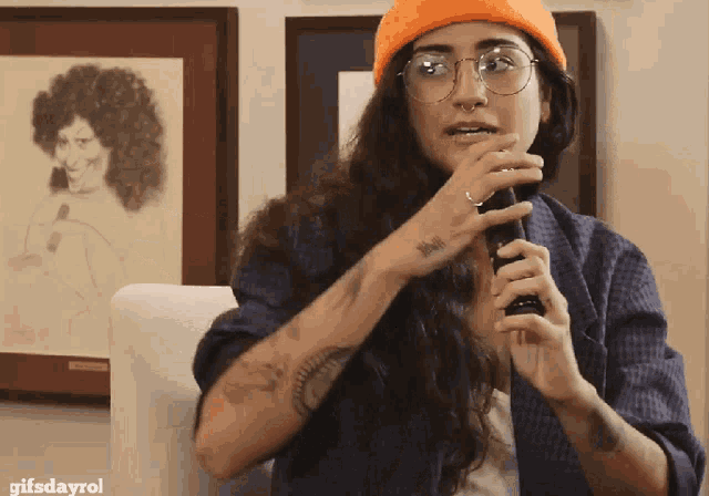 a woman wearing glasses and an orange hat is holding a microphone in front of a framed picture of a woman