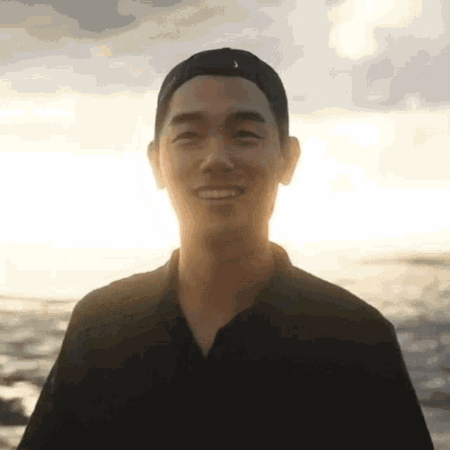 a man wearing a black shirt and a baseball cap is smiling in front of the ocean .