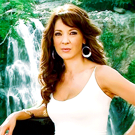 a woman standing in front of a waterfall wearing a white tank top and earrings