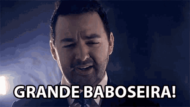 a man in a suit and tie says grande baboseira in a dark room