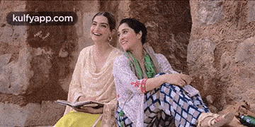 two women are sitting next to each other on a rock . one of the women is holding a book .