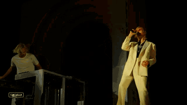 a man in a white suit sings into a microphone while another man plays a keyboard in the background