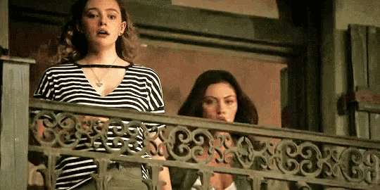 two women are standing on a balcony looking out . one of the women is wearing a striped shirt .
