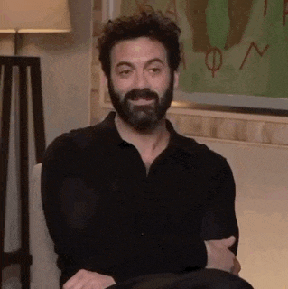 a man with a beard is sitting in a chair with his hands folded and smiling .