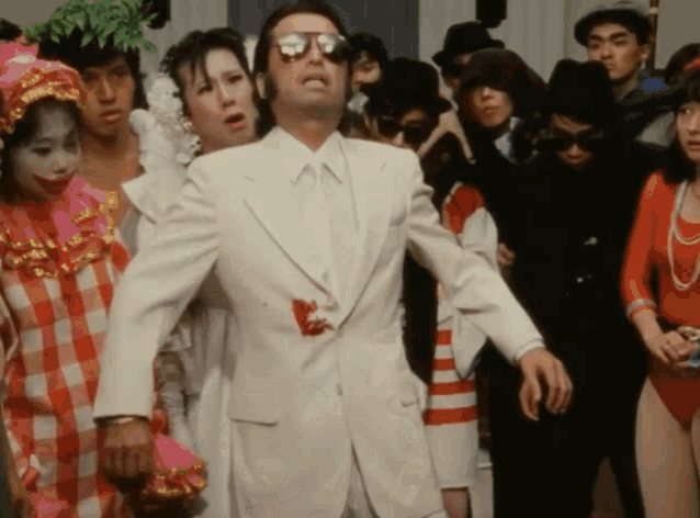 a man in a white suit and sunglasses stands in front of a crowd of people