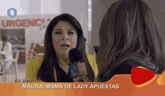 a woman in a yellow jacket is talking into a microphone in front of a sign that says urgencia