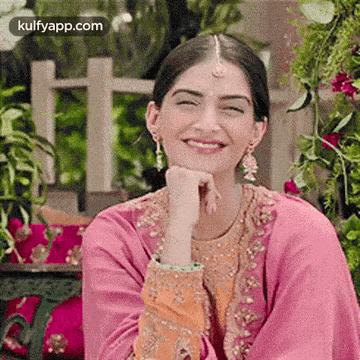 a woman in a pink and orange dress is smiling with her hand under her chin .
