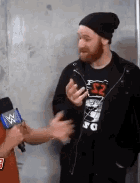 a man with a beard is talking to a reporter while wearing a beanie and a t-shirt .