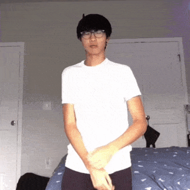 a young man wearing glasses and a white shirt stands in front of a bed
