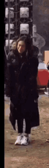 a woman in a black coat and white sneakers is standing on a sidewalk in front of a building .