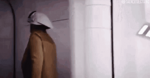 a man in a brown jacket and white helmet is standing in a hallway .