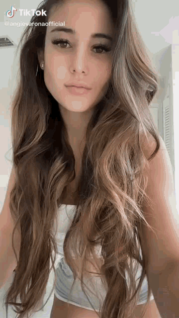 a woman with long hair is wearing a white top and shorts and taking a selfie .