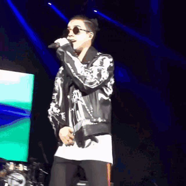 a man wearing sunglasses and a leather jacket is singing into a microphone on stage .