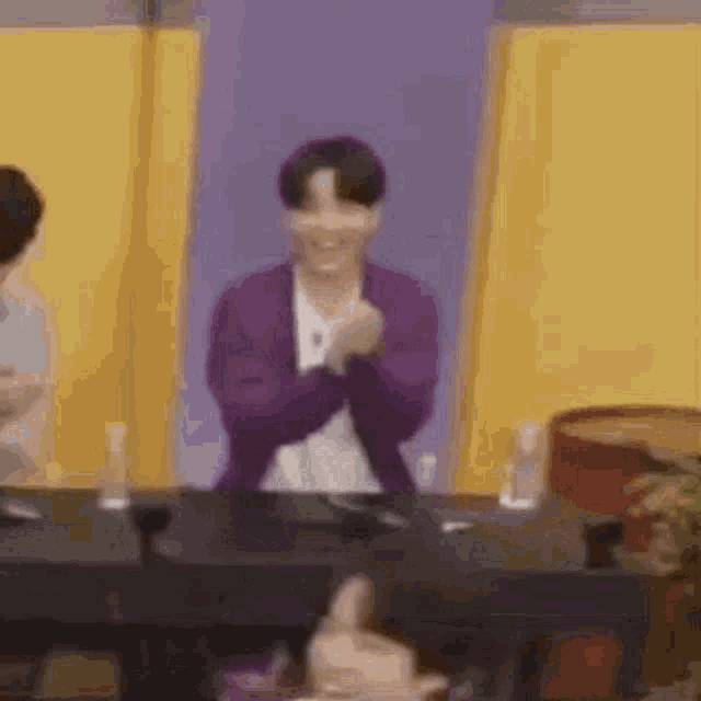 a man in a purple jacket is sitting at a table with his hands in the air .
