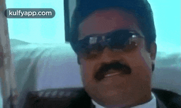 a man with a mustache and sunglasses is sitting in a car and smiling .