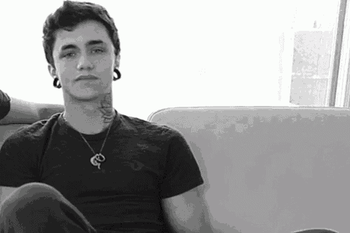 a young man with a tattoo on his neck is sitting on a couch wearing a necklace and earrings .