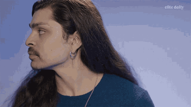 a man with long hair and a mustache is wearing a blue sweater with the elite daily logo behind him