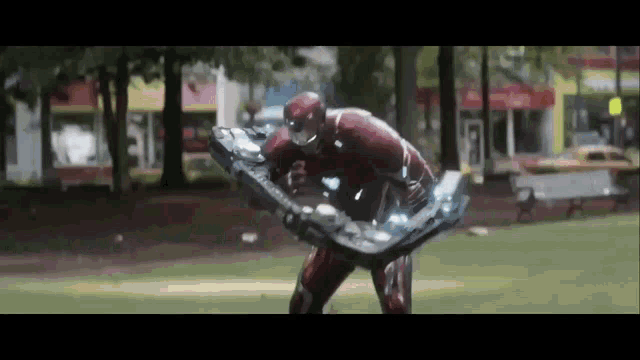 a man in a superhero costume is standing in a park holding a giant shield .