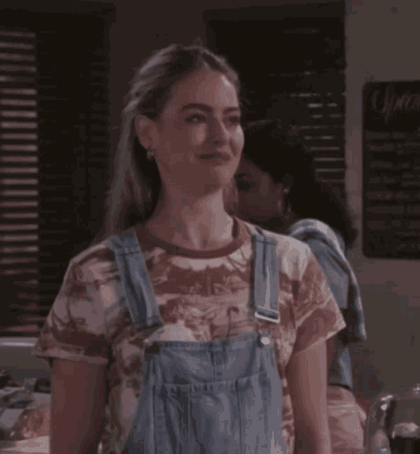 a woman wearing overalls and a tie dye shirt stands in front of a sign that says sprite