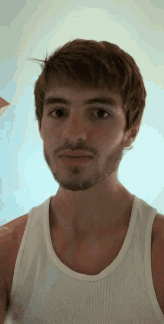 a man with a beard wearing a white tank top looks at the camera
