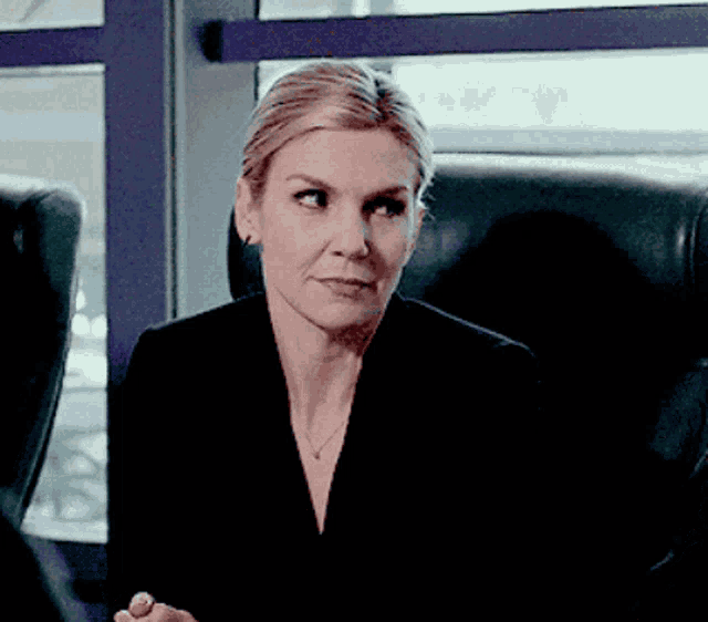 a woman in a black suit is sitting in a chair in front of a window