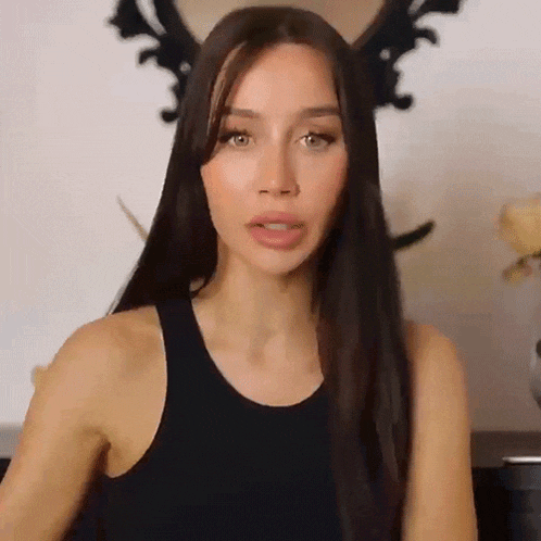 a woman with long brown hair is wearing a black tank top and looking at the camera .