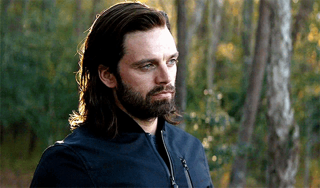 a man with long hair and a beard wearing a black jacket is standing in a forest .