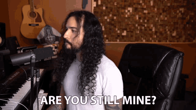 a man with long hair and a beard sitting in front of a microphone with the words are you still mine below him