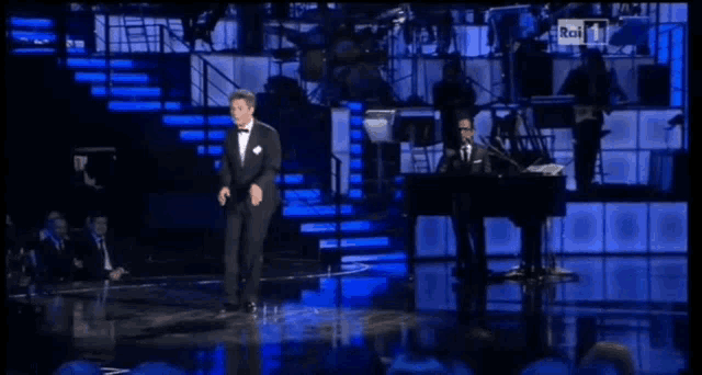 a man in a tuxedo stands on a stage in front of a crowd with rai on the screen