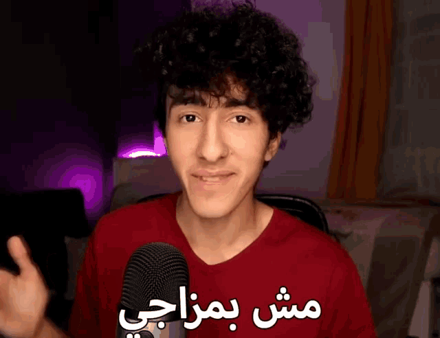 a young man in a red shirt stands in front of a microphone with arabic writing behind him