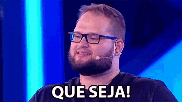 a man with a beard wearing glasses and a microphone says que seja