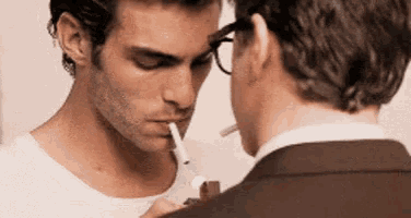 a man with glasses smoking a cigarette next to another man in a suit