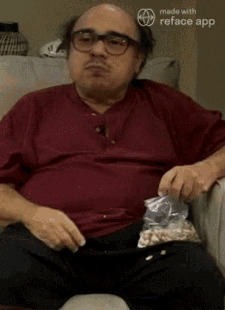 a bald man is sitting on a couch with a bag of popcorn .