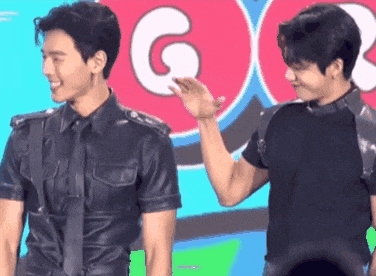 two men are standing next to each other on a stage . one of the men is wearing a leather shirt and tie .