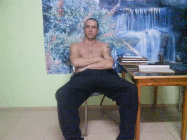 a shirtless man sits at a desk with his arms crossed in front of a waterfall wallpaper