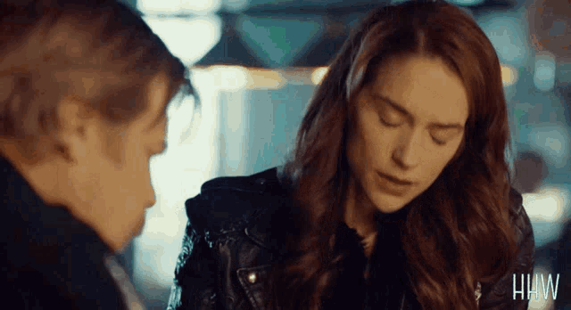 a woman in a leather jacket is talking to a man in a dark room with the letters hw on the bottom left