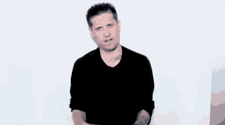 a man in a black shirt is standing in front of a white wall with his hands on his hips .