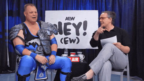 two men sit in front of a sign that says hey ew