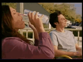 a woman drinking from a bottle while a man sits behind her